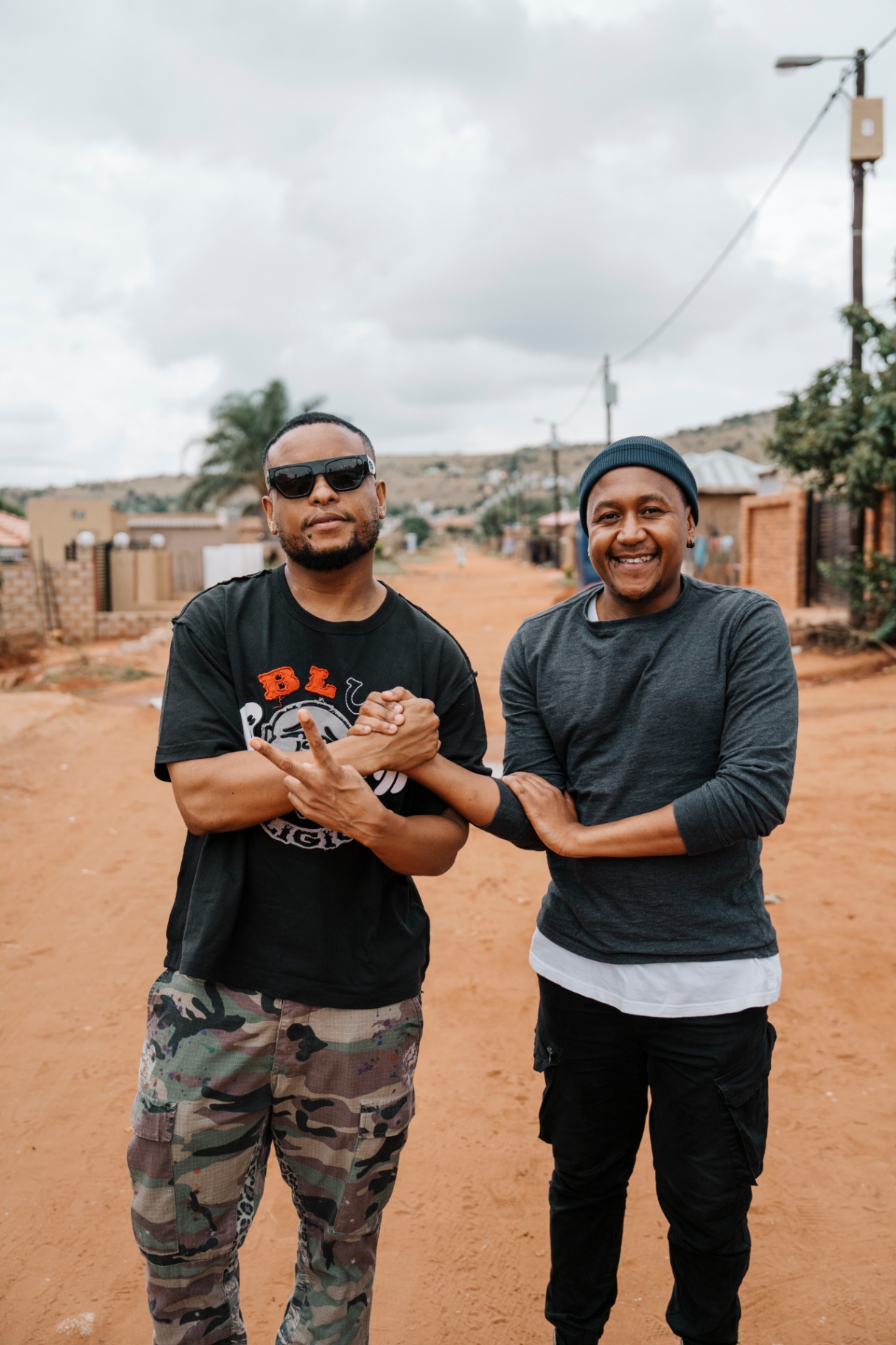Two artists greeting on a dirt road, township homes in the background