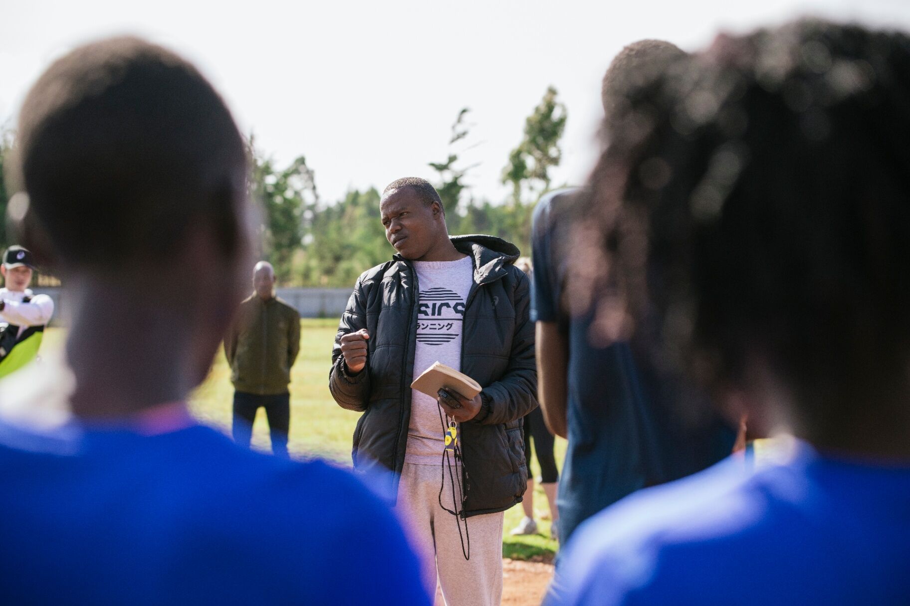 A coach in an ASICS Running tee addresses athletes at trackside in Iten